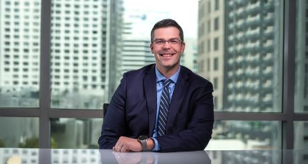 man in suit sitting at a desk with glass windows and buildings in background