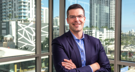 headshot of male in a suit in front of window with office buildings in background